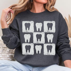 K&ouml;nnte beinhalten: Dunkelgraues Sweatshirt mit einem sich wiederholenden Muster aus wei&szlig;en Z&auml;hnen in quadratischen Rahmen. Das Design hat einen Used-Look. Das Sweatshirt ist ein l&auml;ssiges Langarm-Kleidungsst&uuml;ck, geeignet f&uuml;r Zahn&auml;rzte oder Interessierte der Zahnmedizin.