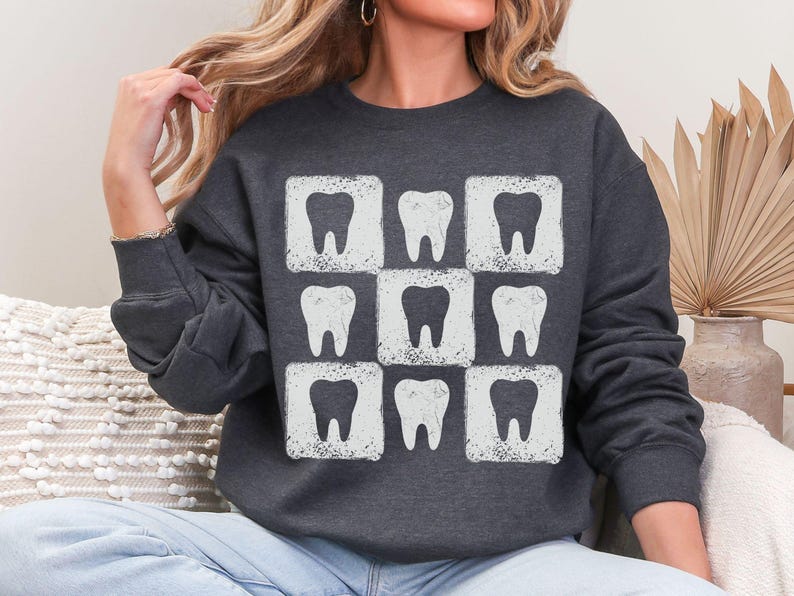 K&ouml;nnte beinhalten: Dunkelgraues Sweatshirt mit einem sich wiederholenden Muster aus wei&szlig;en Z&auml;hnen in quadratischen Rahmen. Das Design hat einen Used-Look. Das Sweatshirt ist ein l&auml;ssiges Langarm-Kleidungsst&uuml;ck, geeignet f&uuml;r Zahn&auml;rzte oder Interessierte der Zahnmedizin.