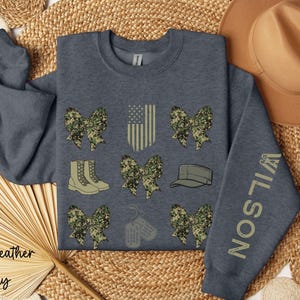 May include: Dark heather gray sweatshirt with military-themed graphics. Features camouflage bows, boots, a cap, dog tags, and an American flag design. The sleeve displays the name "WILSON" in a matching color.