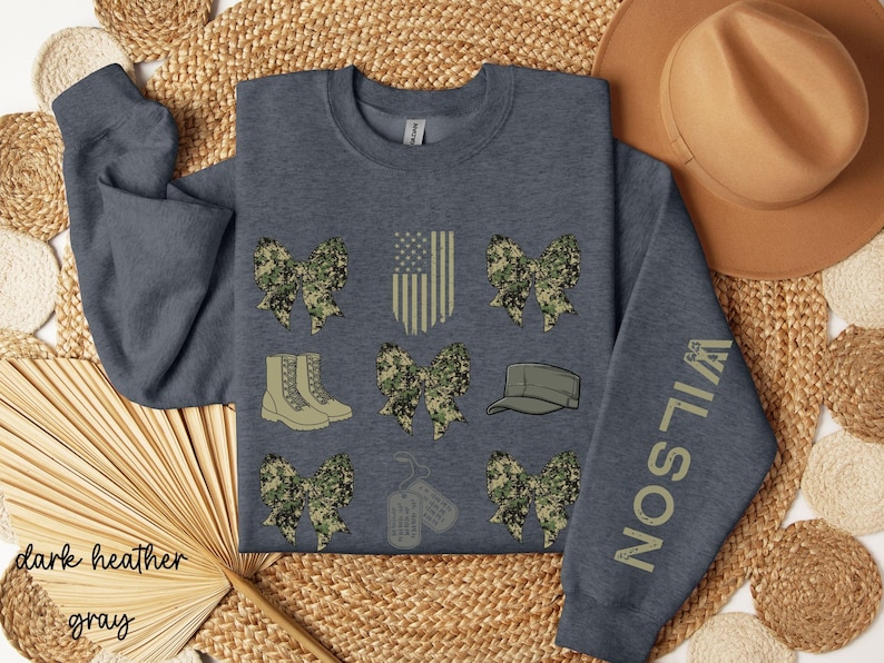 May include: Dark heather gray sweatshirt with military-themed graphics. Features camouflage bows, boots, a cap, dog tags, and an American flag design. The sleeve displays the name "WILSON" in a matching color.
