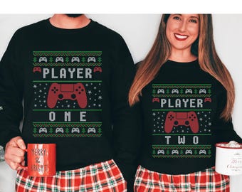 Op de afbeelding: Een paar zwarte kersttruien met een feestelijk ontwerp. De ene trui zegt "Player One" met een rode videogamecontroller en de andere zegt "Player Two" met een rode videogamecontroller.