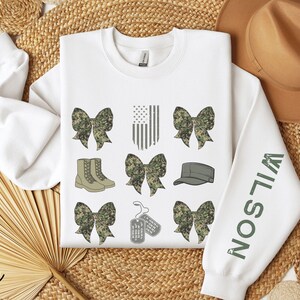May include: White sweatshirt with military-themed graphics. Features camouflage bows, boots, an American flag, a cap, and dog tags. The right sleeve displays the name "WILSON" in green letters. The word "white" is written in the bottom left corner.