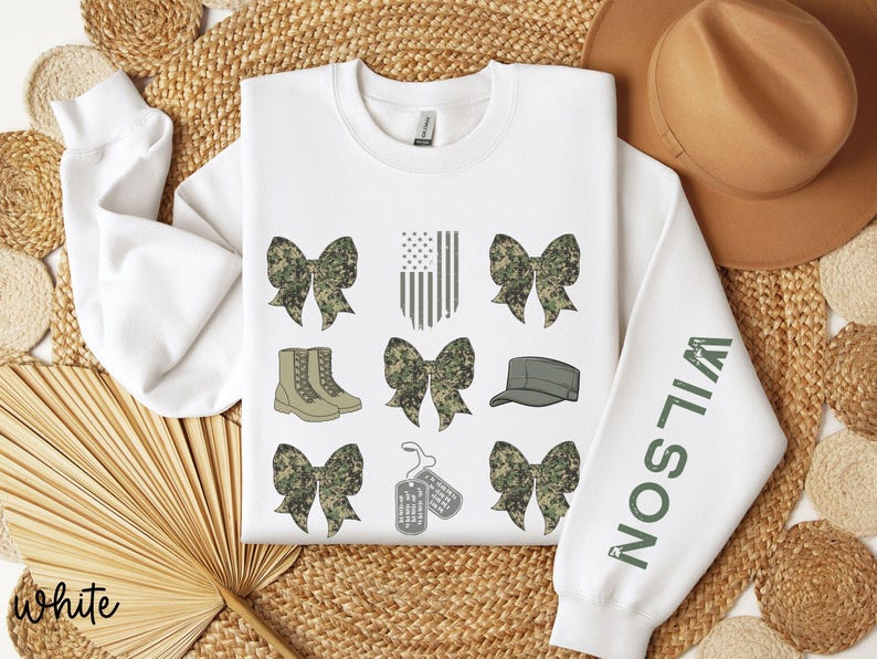May include: White sweatshirt with military-themed graphics. Features camouflage bows, boots, an American flag, a cap, and dog tags. The right sleeve displays the name "WILSON" in green letters. The word "white" is written in the bottom left corner.