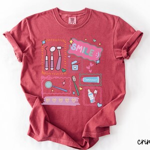 Op de afbeelding: Een karmozijnrood t-shirt met korte mouwen en een grafische print van tandheelkundige instrumenten, waaronder een spiegel, sonde, tandenborstel en tandpasta. Het ontwerp bevat de tekst "SMILE :)" in een roze tekstballon, omringd door hartjes en sterren.