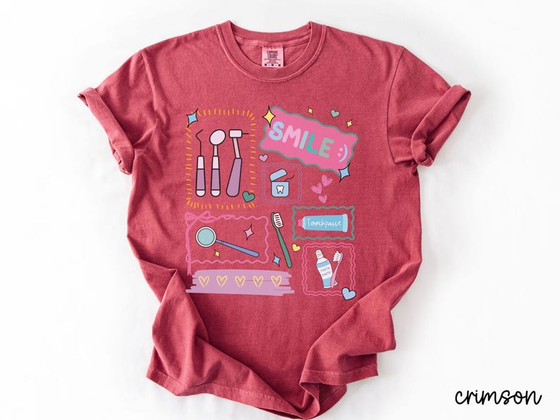 Op de afbeelding: Een karmozijnrood t-shirt met korte mouwen en een grafische print van tandheelkundige instrumenten, waaronder een spiegel, sonde, tandenborstel en tandpasta. Het ontwerp bevat de tekst "SMILE :)" in een roze tekstballon, omringd door hartjes en sterren.