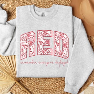 May include: Light gray sweatshirt with the word "RED" in large, distressed red letters filled with a floral pattern. The phrase "remember everyone deployed" is written below in a script font. A casual, comfortable style.