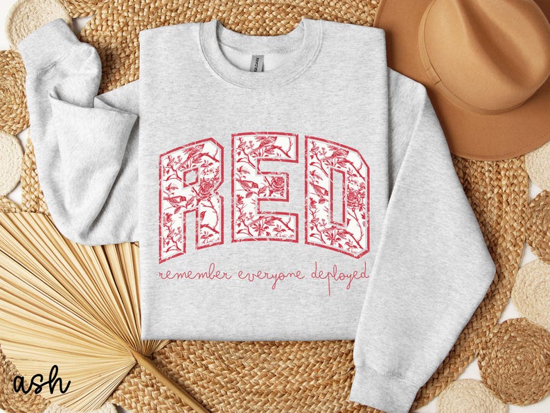 May include: Light gray sweatshirt with the word "RED" in large, distressed red letters filled with a floral pattern. The phrase "remember everyone deployed" is written below in a script font. A casual, comfortable style.