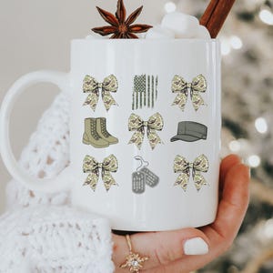 May include: White ceramic mug with a handle, featuring a camouflage pattern design. The design includes bows, boots, a hat, dog tags, and a camouflage American flag.