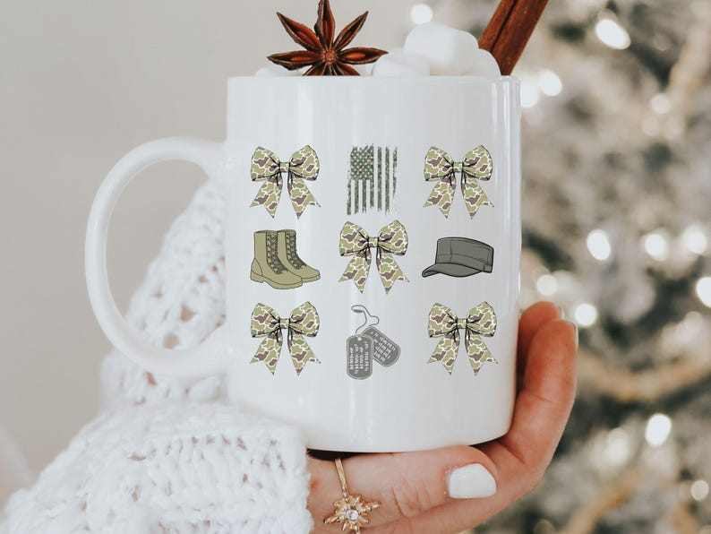 May include: White ceramic mug with a handle, featuring a camouflage pattern design. The design includes bows, boots, a hat, dog tags, and a camouflage American flag.