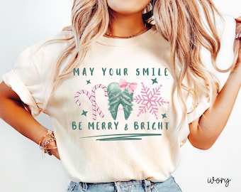 Può includere: Una t-shirt color avorio con una grafica che riporta il testo "MAY YOUR SMILE BE MERRY & BRIGHT" in verde. Il design include un dente verde decorato come un albero di Natale con un fiocco rosa, due bastoncini di zucchero e un fiocco di neve rosa.