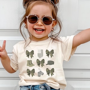 May include: A young child wearing a cream-colored t-shirt with a camouflage print design featuring bows, boots, a hat, and dog tags. The child is wearing sunglasses and has their hair pulled back in a ponytail.