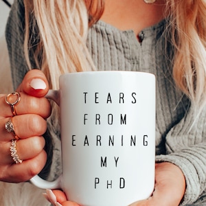 May include: A white ceramic mug with a black handle and the text "But First, Coffee" in a bold, black font. The mug is set against a blurred background, suggesting a cozy setting.