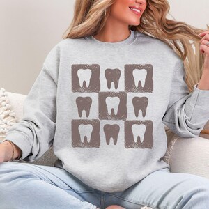 K&ouml;nnte beinhalten: Ein hellgraues Sweatshirt mit einem Muster aus neun braunen Z&auml;hnen in quadratischen Rahmen. Das Design ist ein sich wiederholendes Raster von Zahnillustrationen. Das Sweatshirt ist ein l&auml;ssiges Langarm-Kleidungsst&uuml;ck.