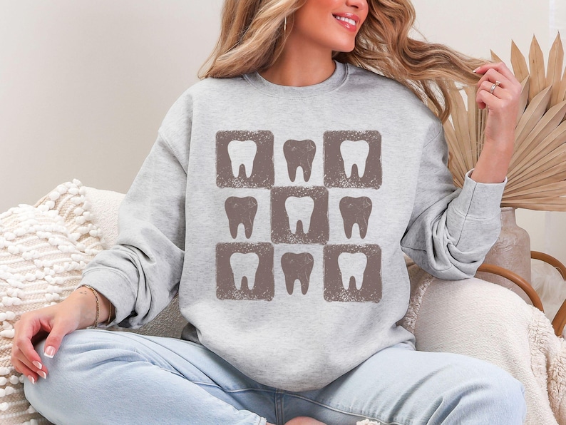 K&ouml;nnte beinhalten: Ein hellgraues Sweatshirt mit einem Muster aus neun braunen Z&auml;hnen in quadratischen Rahmen. Das Design ist ein sich wiederholendes Raster von Zahnillustrationen. Das Sweatshirt ist ein l&auml;ssiges Langarm-Kleidungsst&uuml;ck.