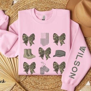 May include: Light pink sweatshirt with military-themed graphics. The design includes camouflage bows, an American flag, combat boots, a military cap, and dog tags. The sleeve features the name "WILSON" in green.