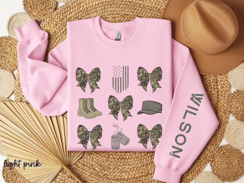 May include: Light pink sweatshirt with military-themed graphics. The design includes camouflage bows, an American flag, combat boots, a military cap, and dog tags. The sleeve features the name "WILSON" in green.