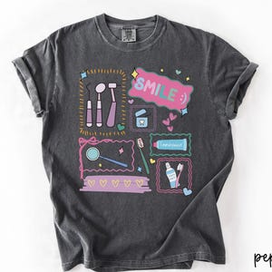Op de afbeelding: Een antracietgrijs t-shirt met korte mouwen en een kleurrijke grafische print van tandheelkundige instrumenten, een tandenborstel, tandpasta en flosdraad. Het ontwerp bevat de tekst "SMILE :)" in een roze tekstballon en kleine hartjes.