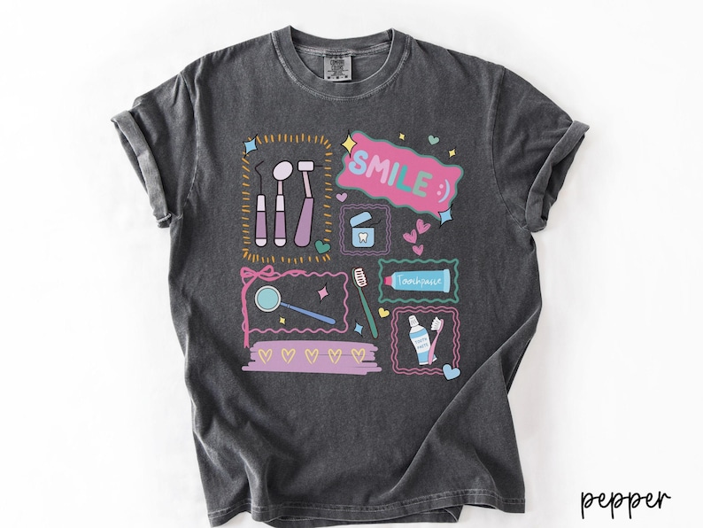 Op de afbeelding: Een antracietgrijs t-shirt met korte mouwen en een kleurrijke grafische print van tandheelkundige instrumenten, een tandenborstel, tandpasta en flosdraad. Het ontwerp bevat de tekst "SMILE :)" in een roze tekstballon en kleine hartjes.