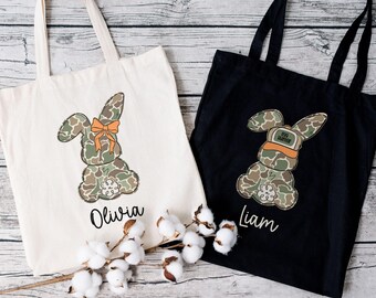 May include: Two personalized tote bags with camouflage bunny designs. One bag is white with an orange bow on the bunny and the name "Olivia". The other bag is black with a camouflage hat on the bunny and the name "Liam".