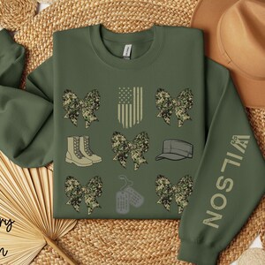 May include: Military green sweatshirt with camouflage bow graphics, an American flag, boots, a cap, and dog tags. The sleeve features the name "WILSON". The words "military green" are at the bottom left.