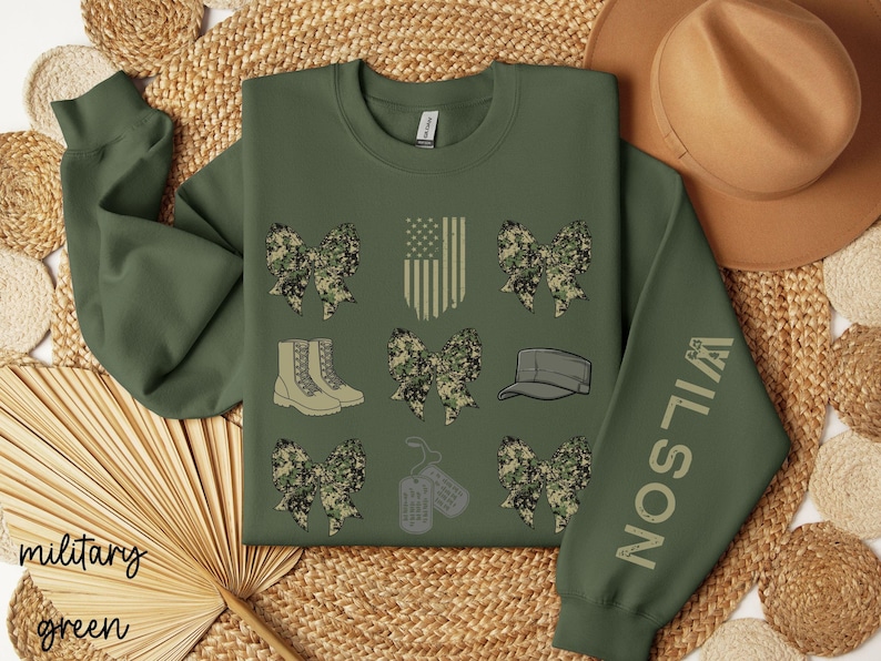May include: Military green sweatshirt with camouflage bow graphics, an American flag, boots, a cap, and dog tags. The sleeve features the name "WILSON". The words "military green" are at the bottom left.