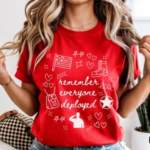 May include: Red t-shirt with white graphic text and illustrations. The text reads "remember everyone deployed" with images of a flag, stars, hearts, a boot, and a military dog tag. The word "red" is printed at the bottom right.