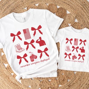 May include: White t-shirts with red graphics, including bows, an American flag, military boots, and silhouettes. The text "remember everyone deployed" is printed on the shirts. The shirts are displayed on a woven mat.