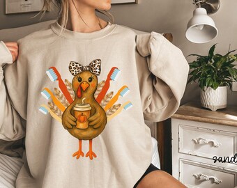 Può includere: Felpa beige con un tacchino dei cartoni animati che tiene in mano un pumpkin spice latte. Il tacchino ha un fiocco ed è circondato da spazzolini da denti. Il design è nei toni del marrone, arancione e blu. La parola "sand" è scritta in corsivo in basso a destra.