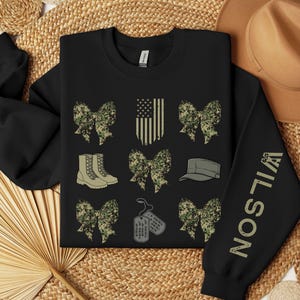 May include: Black sweatshirt with military-themed graphics. Features camouflage bows, boots, a cap, dog tags, and an American flag design. The right sleeve displays the name "WILSON" in a vertical font. The word "black" is at the bottom left.