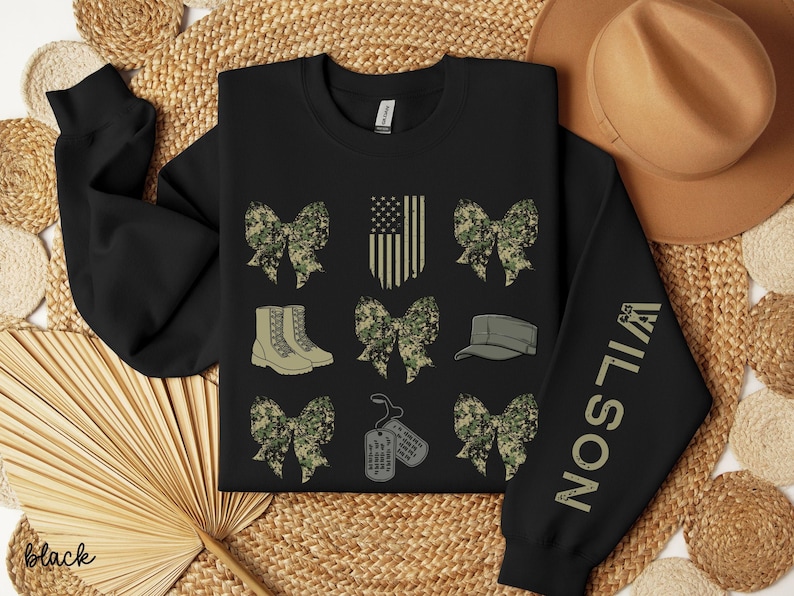 May include: Black sweatshirt with military-themed graphics. Features camouflage bows, boots, a cap, dog tags, and an American flag design. The right sleeve displays the name "WILSON" in a vertical font. The word "black" is at the bottom left.
