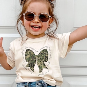 Peut inclure: Un t-shirt blanc avec un n&oelig;ud en camouflage vert et le texte "Un h&eacute;ros a mon c&oelig;ur" r&eacute;p&eacute;t&eacute; autour du n&oelig;ud.