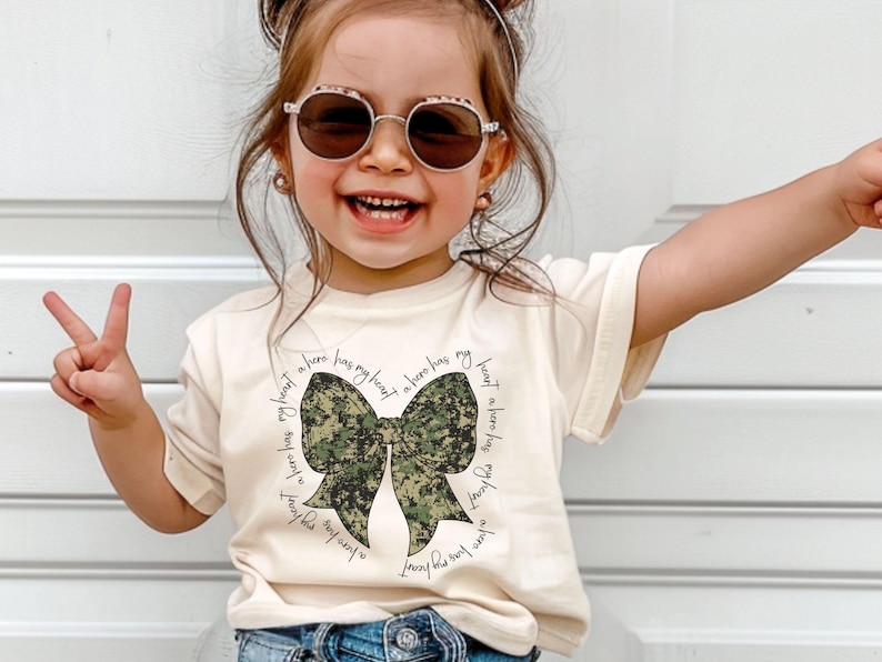 Peut inclure: Un t-shirt blanc avec un n&oelig;ud en camouflage vert et le texte "Un h&eacute;ros a mon c&oelig;ur" r&eacute;p&eacute;t&eacute; autour du n&oelig;ud.
