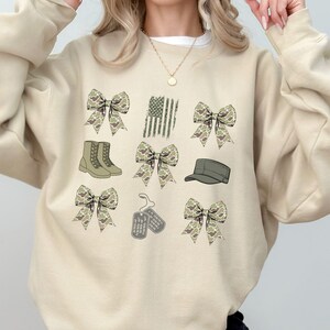 May include: A beige sweatshirt with a camouflage pattern. The sweatshirt features a variety of images including bows, boots, a hat, dog tags, and an American flag.