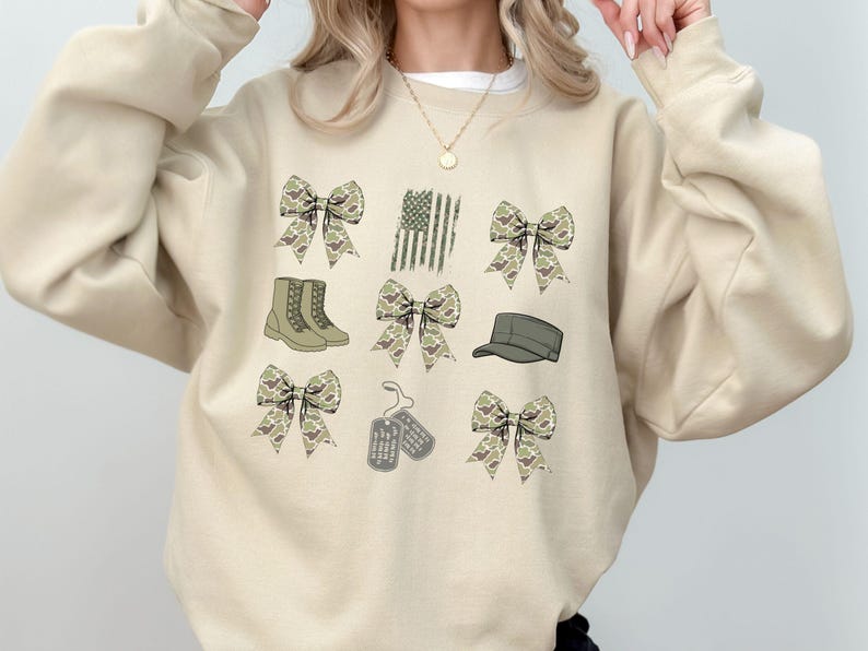 May include: A beige sweatshirt with a camouflage pattern. The sweatshirt features a variety of images including bows, boots, a hat, dog tags, and an American flag.