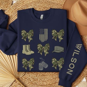 May include: Navy blue sweatshirt with military-themed graphics. Features camouflage bows, combat boots, a military cap, dog tags, and an American flag design. The sleeve displays the name "WILSON" in a vertical font.