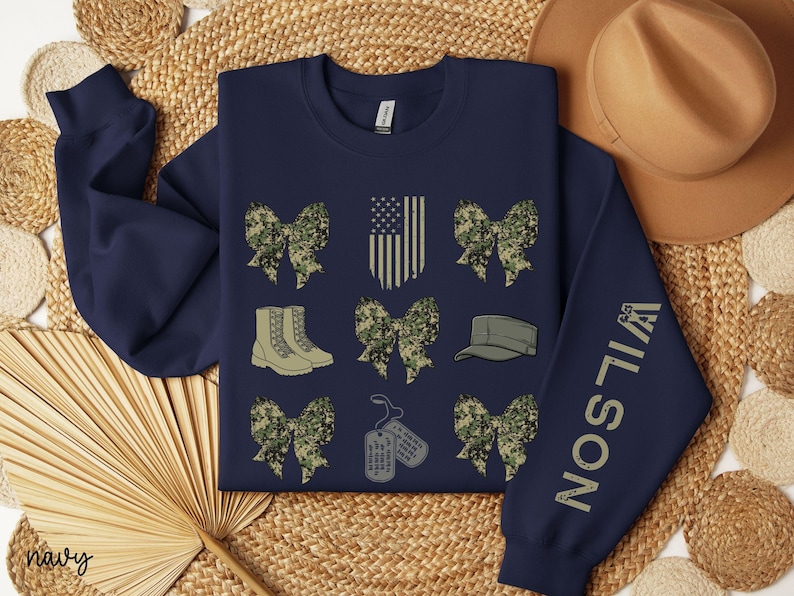May include: Navy blue sweatshirt with military-themed graphics. Features camouflage bows, combat boots, a military cap, dog tags, and an American flag design. The sleeve displays the name "WILSON" in a vertical font.