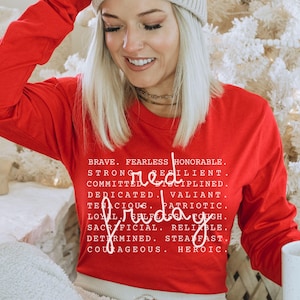 May include: A bright red long-sleeve shirt with the words "Red Friday" and a list of positive attributes printed in white. The shirt is worn by a person with blonde hair and a white beanie.