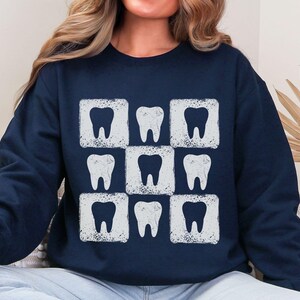 K&ouml;nnte beinhalten: Marineblaues Sweatshirt mit einem sich wiederholenden Muster aus neun wei&szlig;en Zahngrafiken. Jeder Zahn befindet sich in einem wei&szlig;en Quadrat, wodurch ein Raster entsteht. Das Design hat einen leicht verwitterten, Vintage-Look. Das Sweatshirt ist ein l&auml;ssiges, bequemes Kleidungsst&uuml;ck.