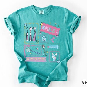 Op de afbeelding: Een zeegroen t-shirt met korte mouwen en een grafische print van tandheelkundige instrumenten, een tube tandpasta, een tandenborstel en flosdraad. Het ontwerp bevat de tekst "SMILE :)" in een roze tekstballon, omringd door kleine hartjes en sterren.