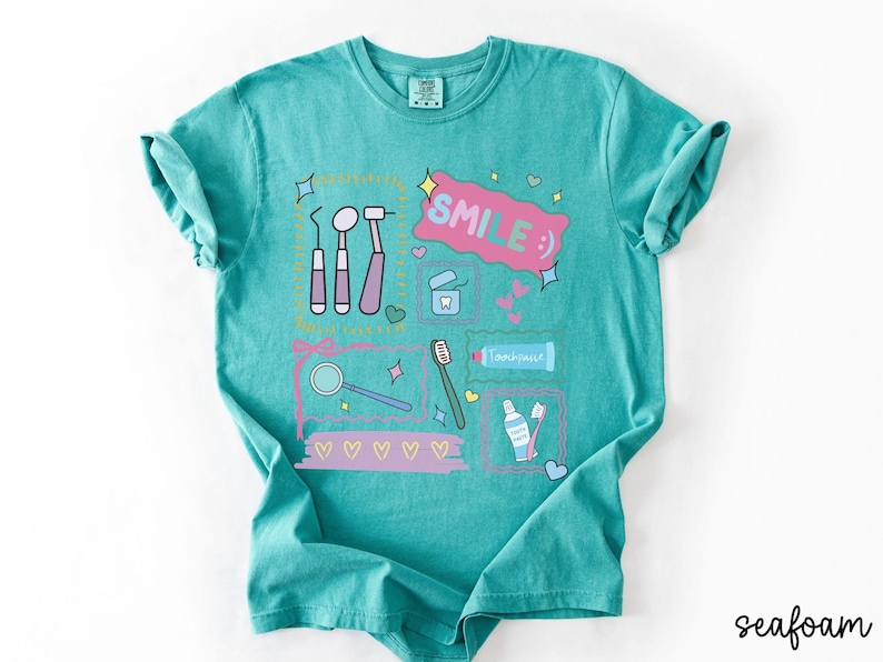 Op de afbeelding: Een zeegroen t-shirt met korte mouwen en een grafische print van tandheelkundige instrumenten, een tube tandpasta, een tandenborstel en flosdraad. Het ontwerp bevat de tekst "SMILE :)" in een roze tekstballon, omringd door kleine hartjes en sterren.