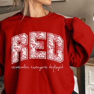May include: A red sweatshirt with the word "RED" in large, white, floral-patterned letters. Below the word "RED" is the phrase "remember everyone deployed" in a cursive font. The sweatshirt is a solid red color.