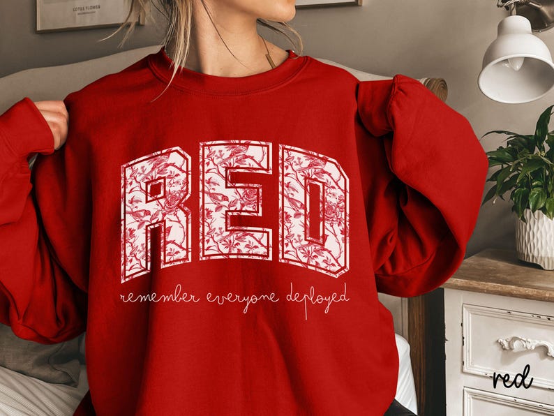 May include: A red sweatshirt with the word "RED" in large, white, floral-patterned letters. Below the word "RED" is the phrase "remember everyone deployed" in a cursive font. The sweatshirt is a solid red color.
