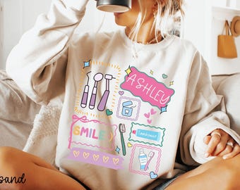 Può includere: Una felpa color sabbia con grafica a tema dentale. Il design include strumenti odontoiatrici, uno spazzolino da denti, dentifricio e il nome "ASHLEY" in una bolla rosa. Viene visualizzata anche la parola "SMILE".