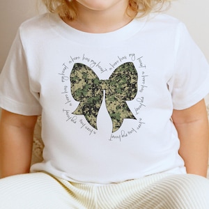 Peut inclure: T-shirt blanc avec un n&oelig;ud camouflage et le texte "My heart has a hero has my heart a hero has my heart a hero has my heart".