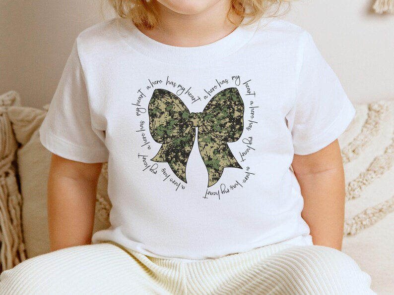 Peut inclure: T-shirt blanc avec un n&oelig;ud camouflage et le texte "My heart has a hero has my heart a hero has my heart a hero has my heart".