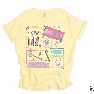 Op de afbeelding: Een lichtgeel t-shirt met een kleurrijke grafische print van tandheelkundige instrumenten, waaronder een spiegel, sonde, tandenborstel en tandpasta. Het ontwerp bevat de tekst "SMILE :)" in een roze tekstballon, omringd door kleine hartjes en sterren.