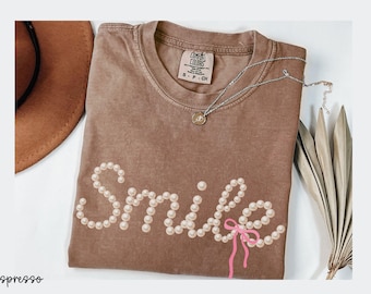 Può includere: Una maglietta marrone con la parola "Smile" scritta con perle finte e un fiocco rosa. Sono visibili anche una collana d'argento con un ciondolo e un cappello marrone. La maglietta è etichettata "espresso" in basso a sinistra.