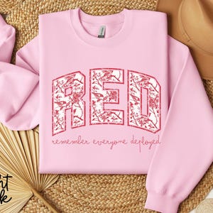 May include: Light pink sweatshirt with the word "RED" in a distressed, floral-filled design. The text "remember everyone deployed" is below the word "RED". The sweatshirt is displayed on a woven surface.