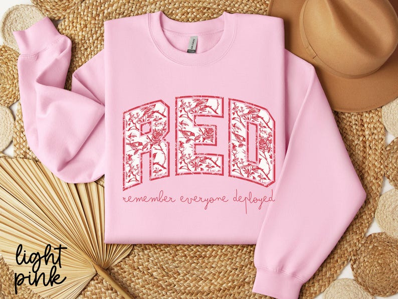 May include: Light pink sweatshirt with the word "RED" in a distressed, floral-filled design. The text "remember everyone deployed" is below the word "RED". The sweatshirt is displayed on a woven surface.