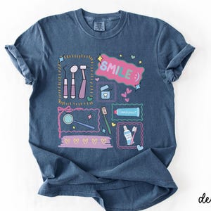 Op de afbeelding: Een denimblauw t-shirt met een grafische print van tandheelkundige instrumenten, een tandenborstel, tandpasta en flosdraad. Het ontwerp bevat de tekst "SMILE :)" in een roze tekstballon, omringd door kleine hartjes, sterretjes en decoratieve kaders.